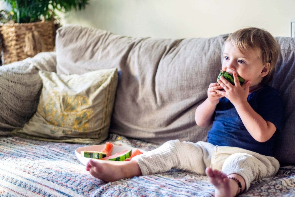 A two year old sat on a grey sofa enthusiastically tucking in to some sliced watermelon.