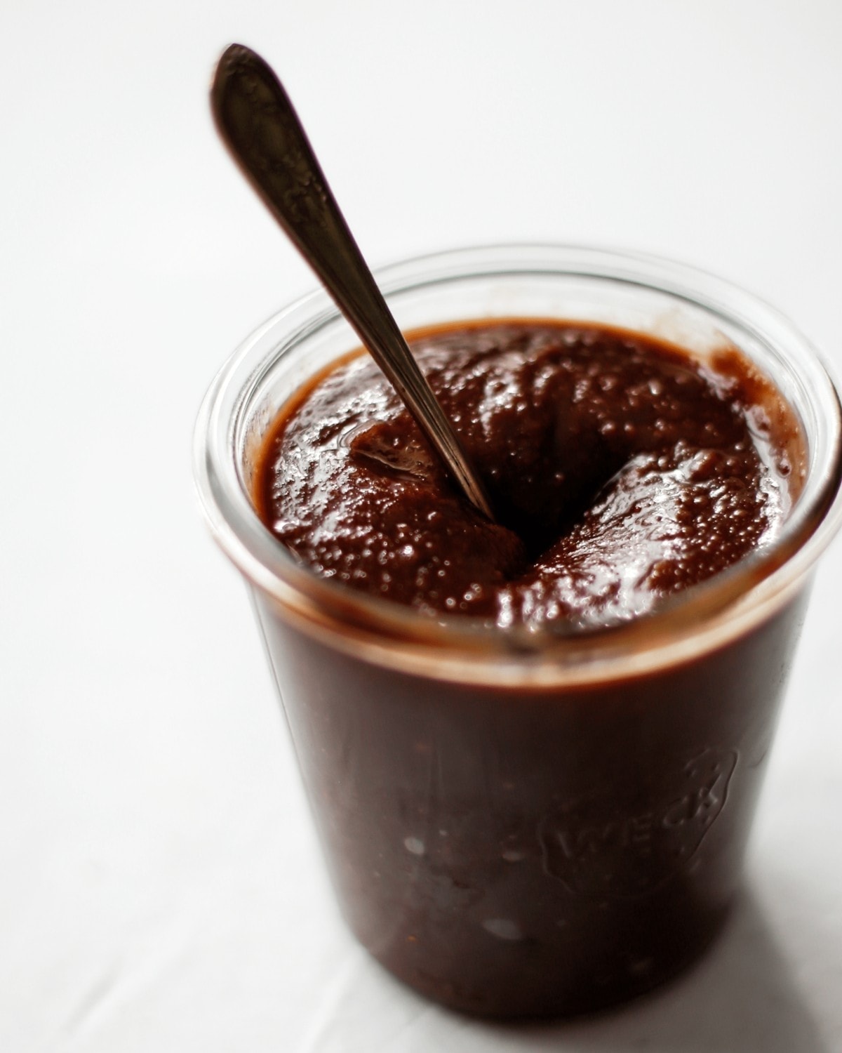 Side view of the chocolate orange spread in a glass jar, with a spoon stood in the middle, after being stirred.