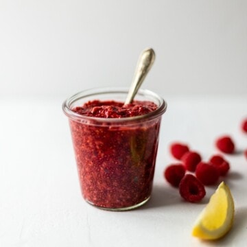 Jar of chia raspberry jam with a spoon in and some raspberries and a lemon wedge scattered next to it.