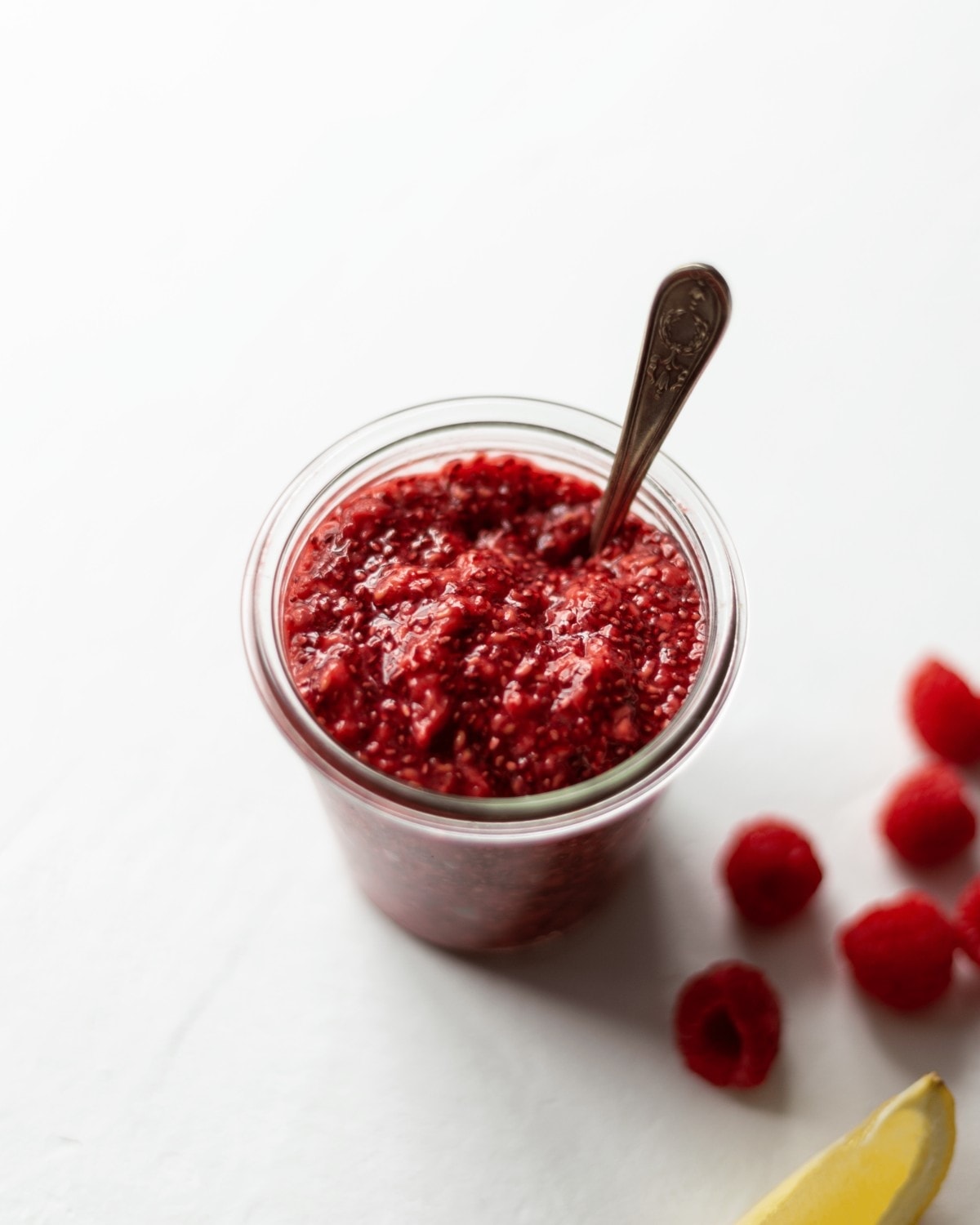 A jar of vegan raspberry chia jam with a spoon in and some raspberries and a lemon wedge scattered next to it.