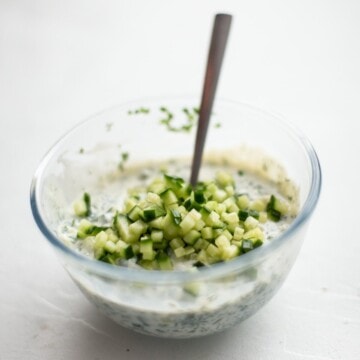 Vegan tzatziki being mixed in a glass bowl, with a spoon. With some diced cucumber on the top before being stirred in.