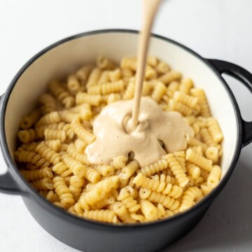 Cheesy cashew sauce being poured over cooked pasta in a black casserole dish, before baking.