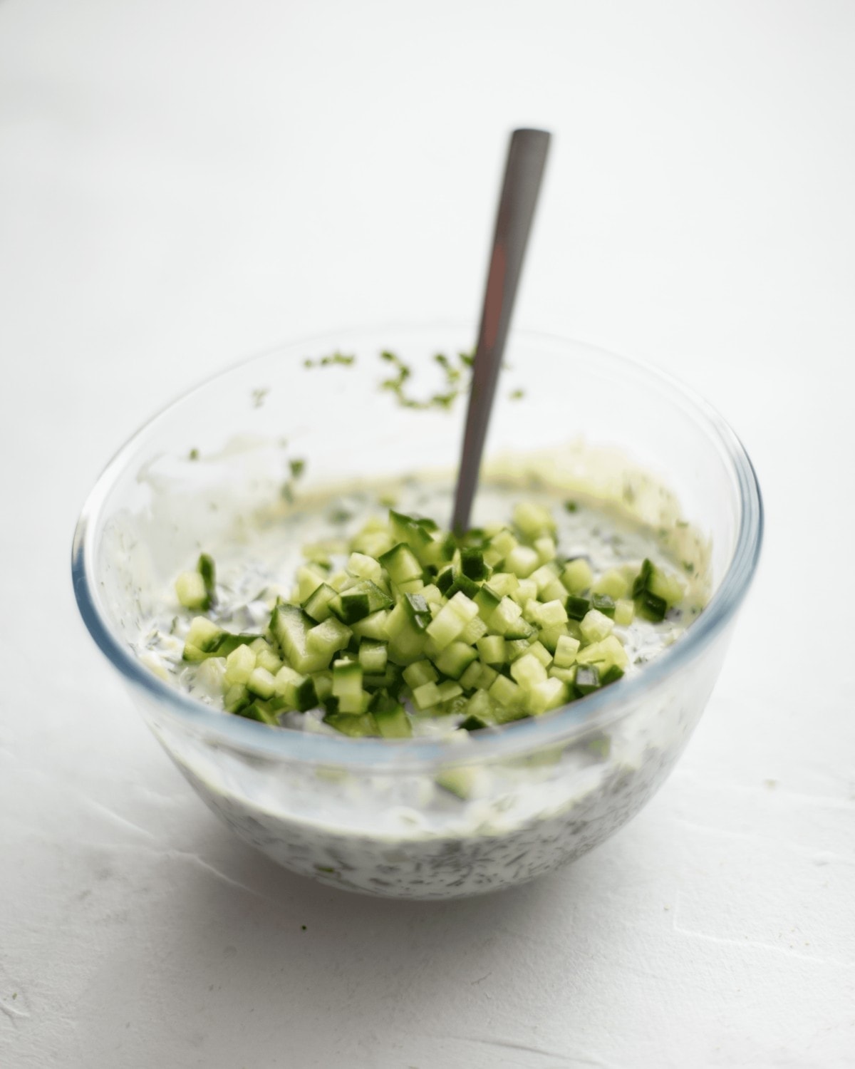 Vegan tzatziki being mixed in a glass bowl, with a spoon. With some diced cucumber on the top before being stirred in.