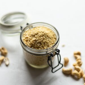 A jar of the vegan parmesan sat on a table with some cashews scattered around.