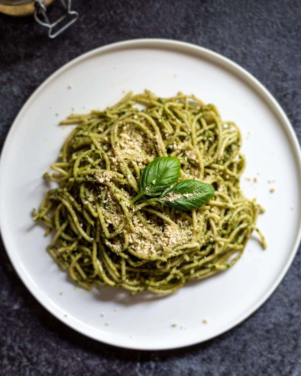 A white plate with wild garlic and pine nut coated spaghetti, topped with vegan parmesan and fresh basil leaves.