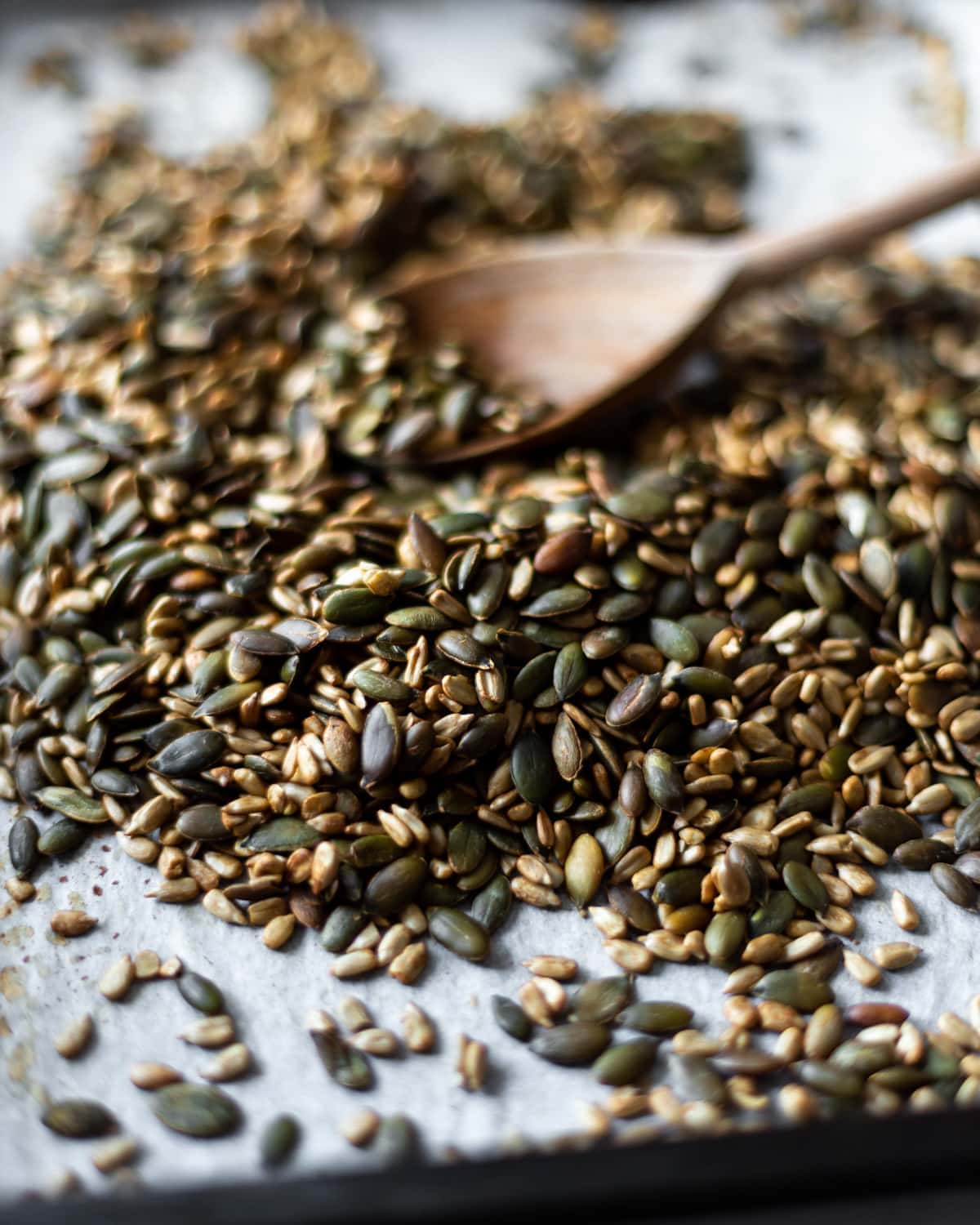 Side shot of the roasted tamari seeds, being stirred through with a wooden spoon, on the baking tray after roasting.