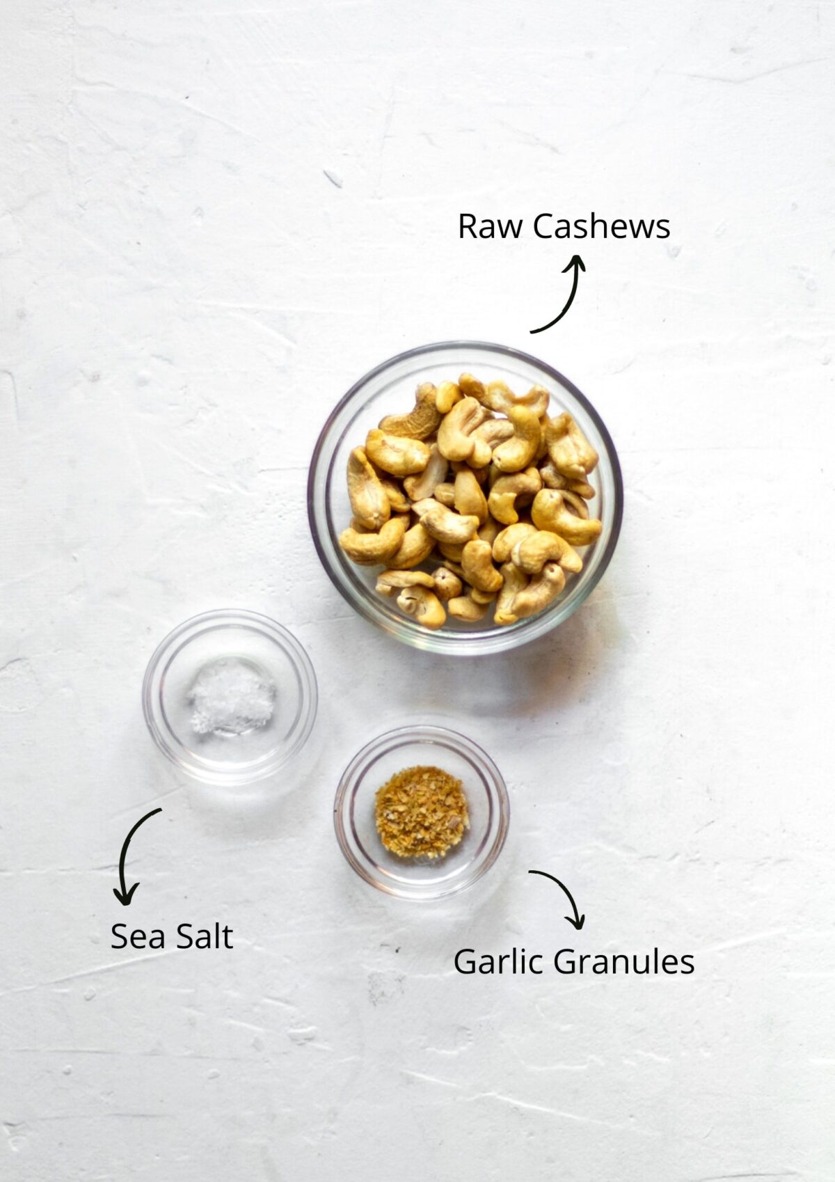 Overhead photo of the ingredients for garlic cashew cream, in glass bowls, on a white background.