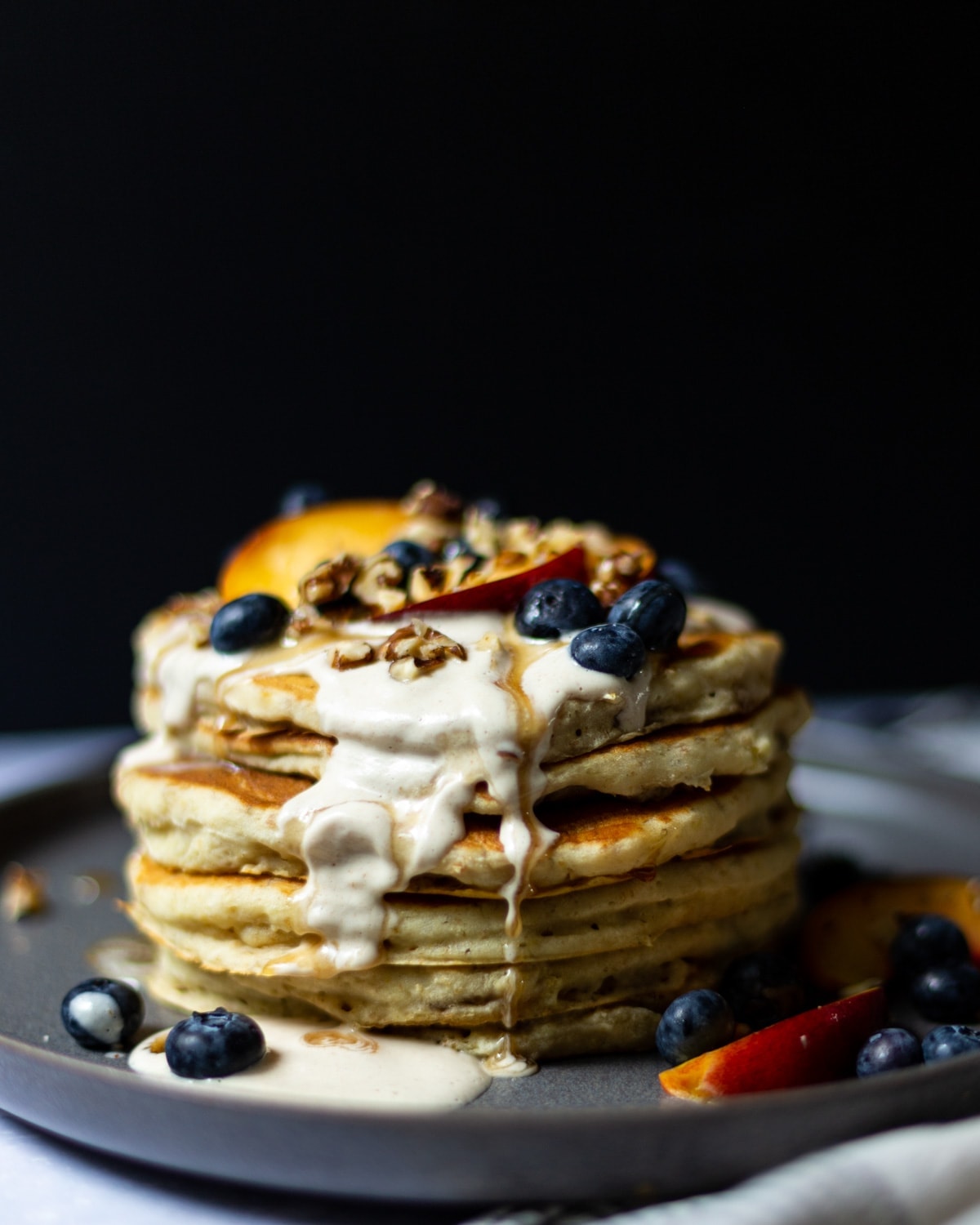 Side view of a stack of pancakes topped with cashew cream, sliced nectarines, blueberries, chopped walnuts and maple syrup.