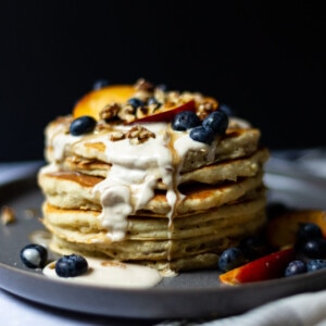 A stack of gluten free pancakes, drizzled with cashew cream, maple syrup and topped with sliced nectarine, fresh blueberries and chopped walnuts.