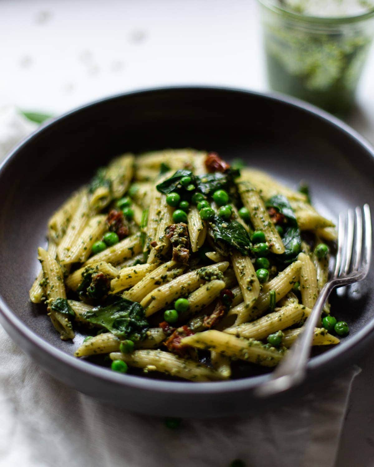 A side-on photo of pesto Penne pasta in a dark grey bowl, with a silver fork in the bowl on the right hand side, and the jar of fresh pesto in the background.