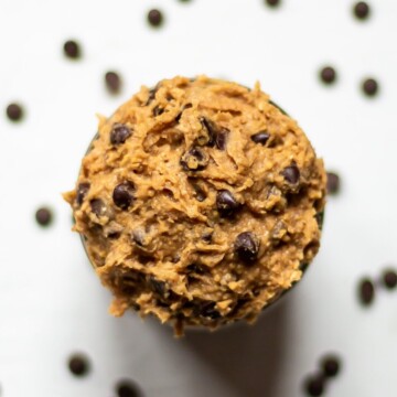 Overhead image of a bowl of chocolate chip edible vegan cookie dough, with more chocolate chips, out of focus, scattered in the background.