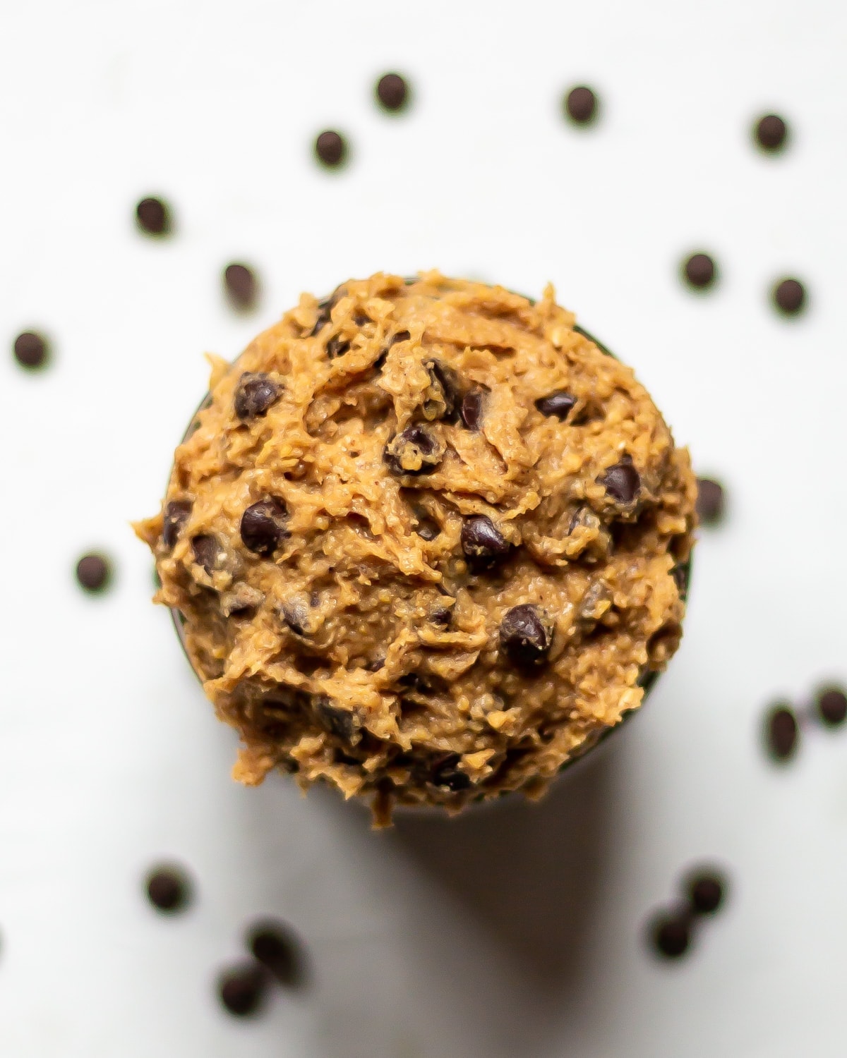 Overhead image of a bowl of chocolate chip edible vegan cookie dough, with more chocolate chips, out of focus, scattered in the background.