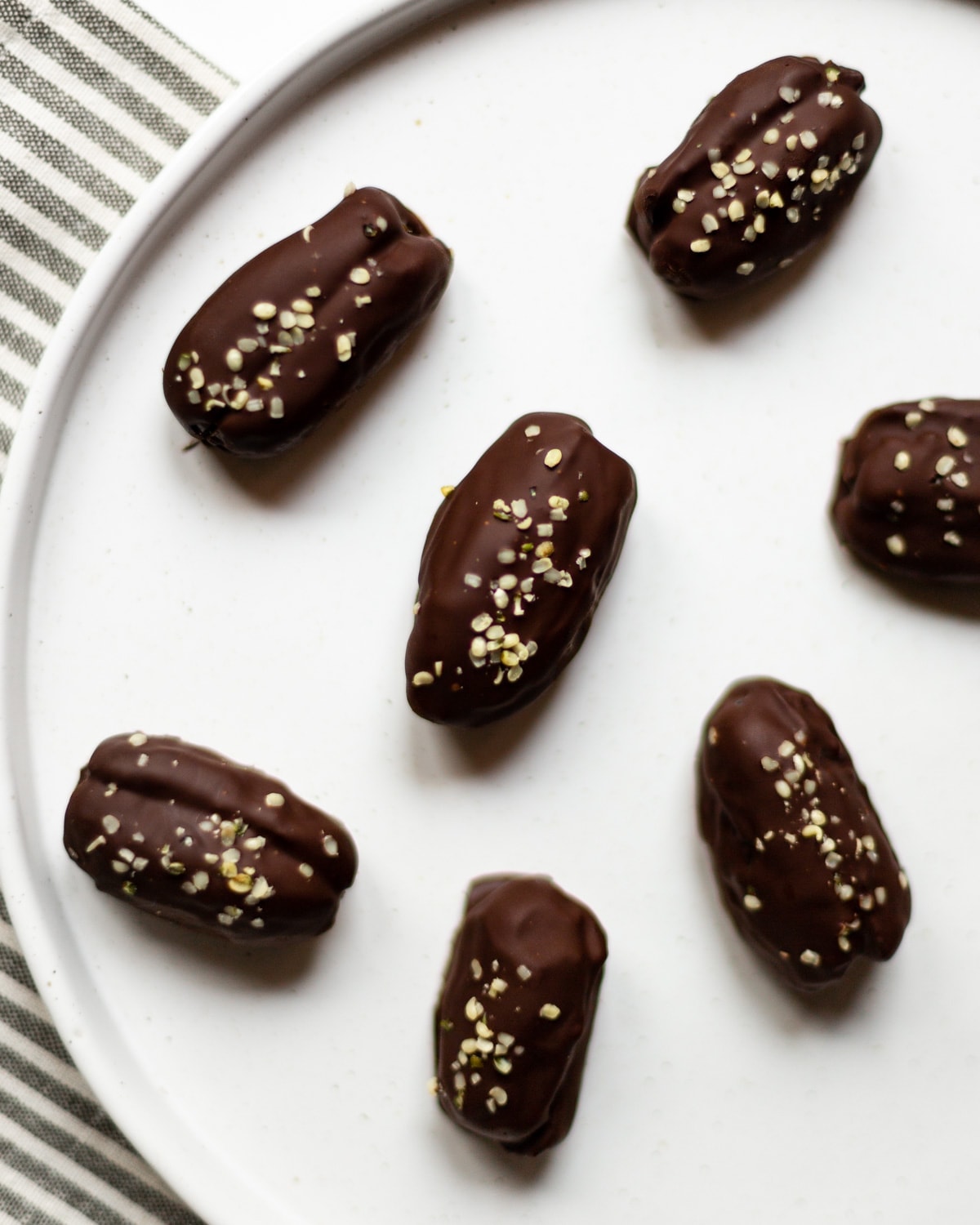 Chocolate coated peanut butter stuffed dates topped with hemp seeds, on a white plate, with a grey striped linen underneath in the background.
