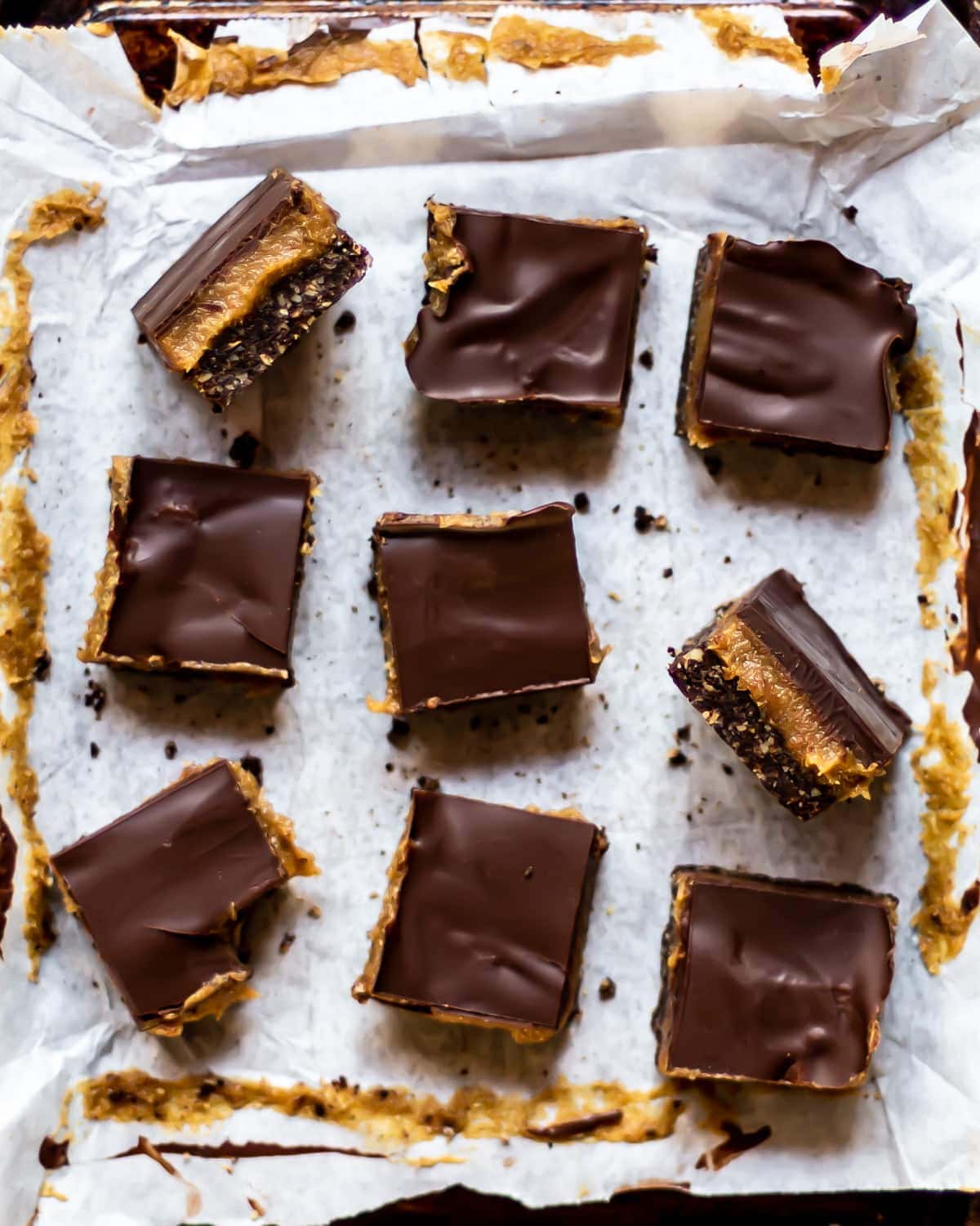9 vegan caramel slices cut in to squares, on a white sheet of greaseproof paper. Two slices have been turned on their side to show the layers within - the base, caramel filling, and chocolate topping.