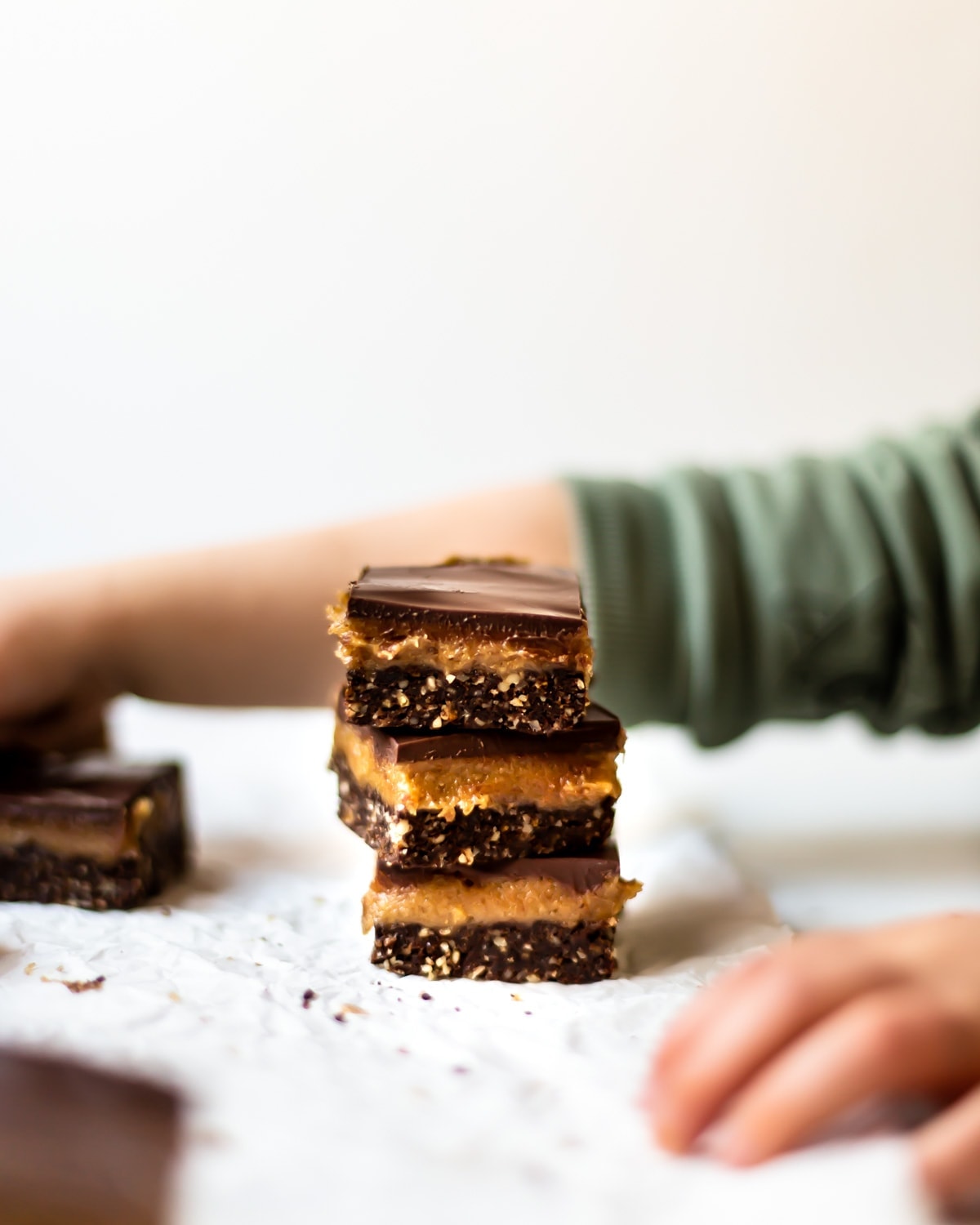 A stack of vegan caramel slices with a toddlers hand in the foreground, about to help himself to one.