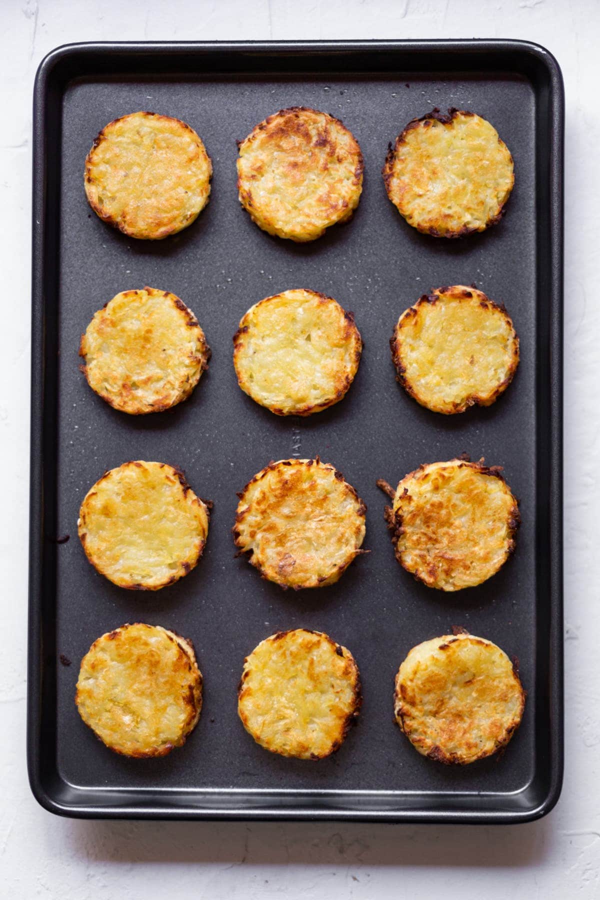 A black baking tray filled with 12 golden baked hash browns.