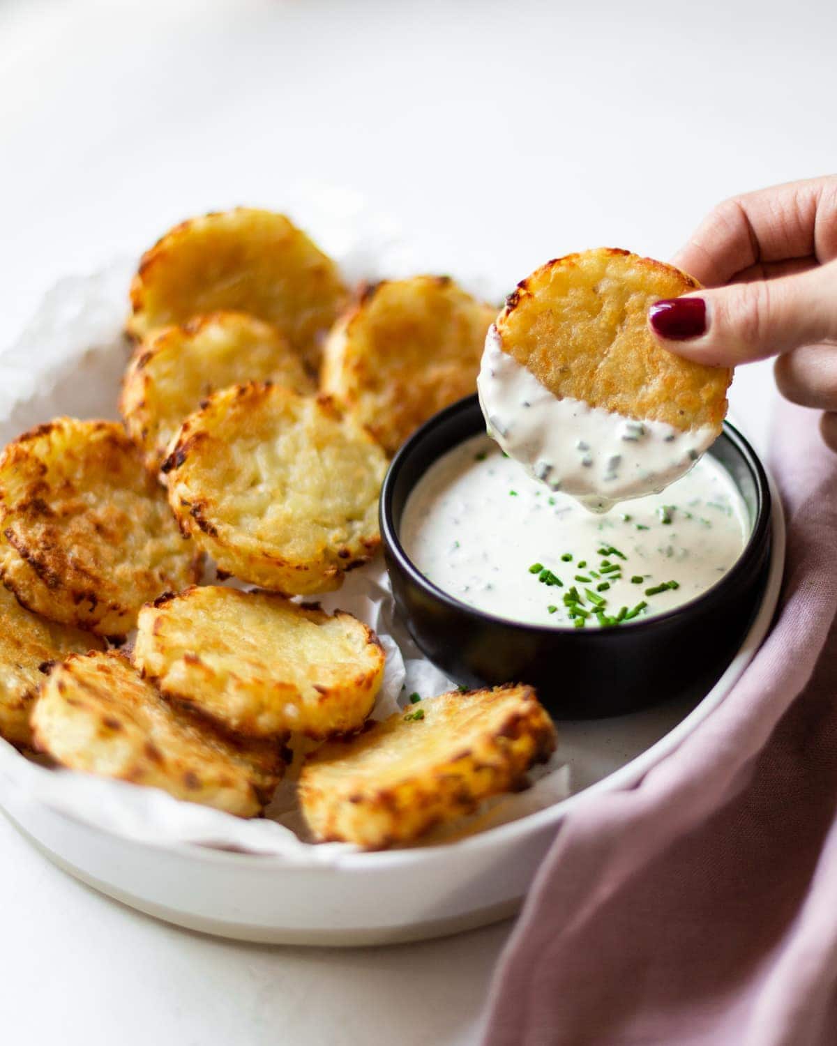 Side angle of a white bowl filled with the baked hash browns, and a black bowl filled with chive dip. One hash brown is being dipped in the chive dip.