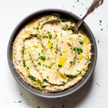 A dark grey bowl, filled with hummus, on a white background. The hummus has been drizzled with olive oil and fresh chopped parsley is sprinkled over.