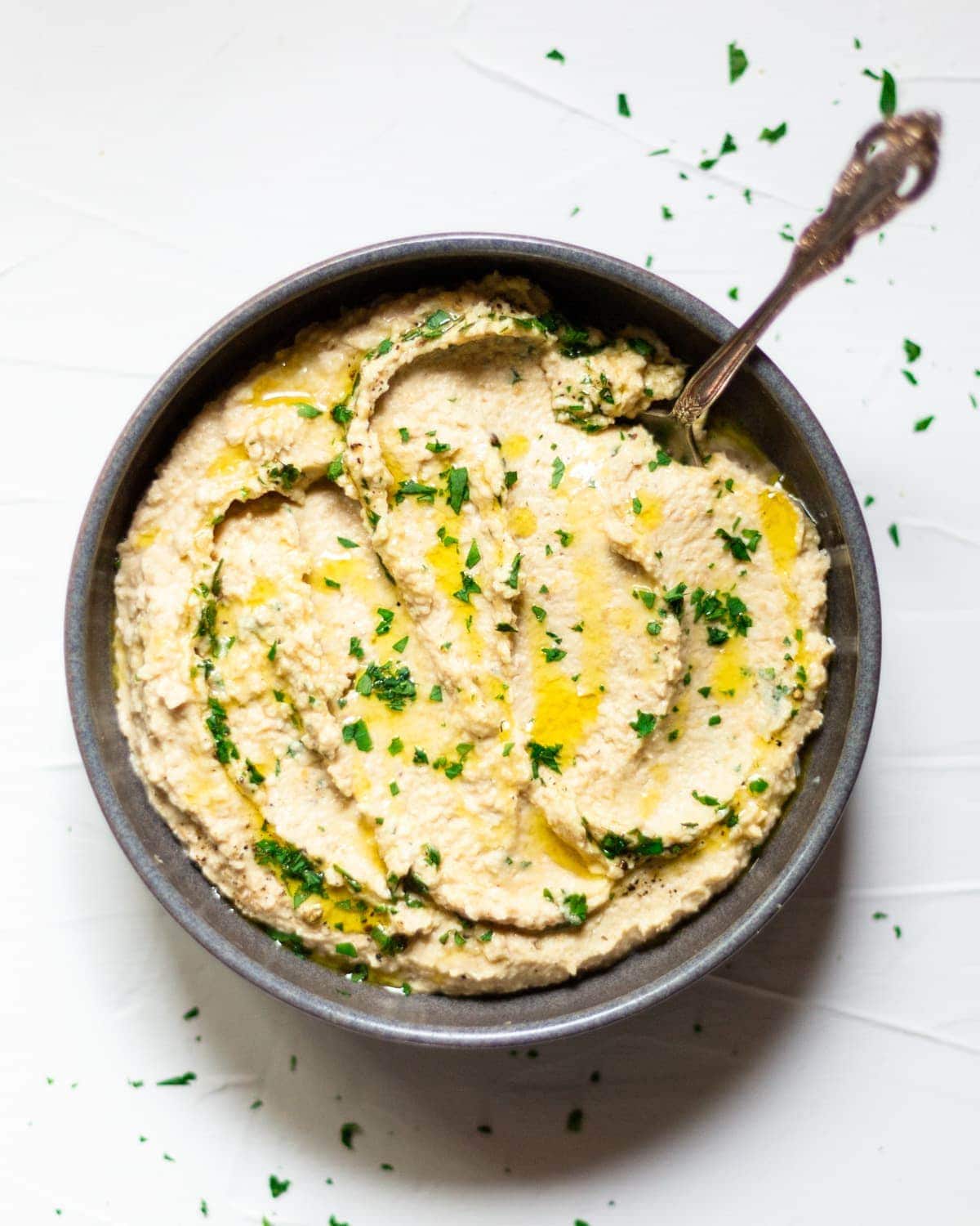 A dark grey bowl, filled with hummus, on a white background. The hummus has been drizzled with olive oil and fresh chopped parsley is sprinkled over.