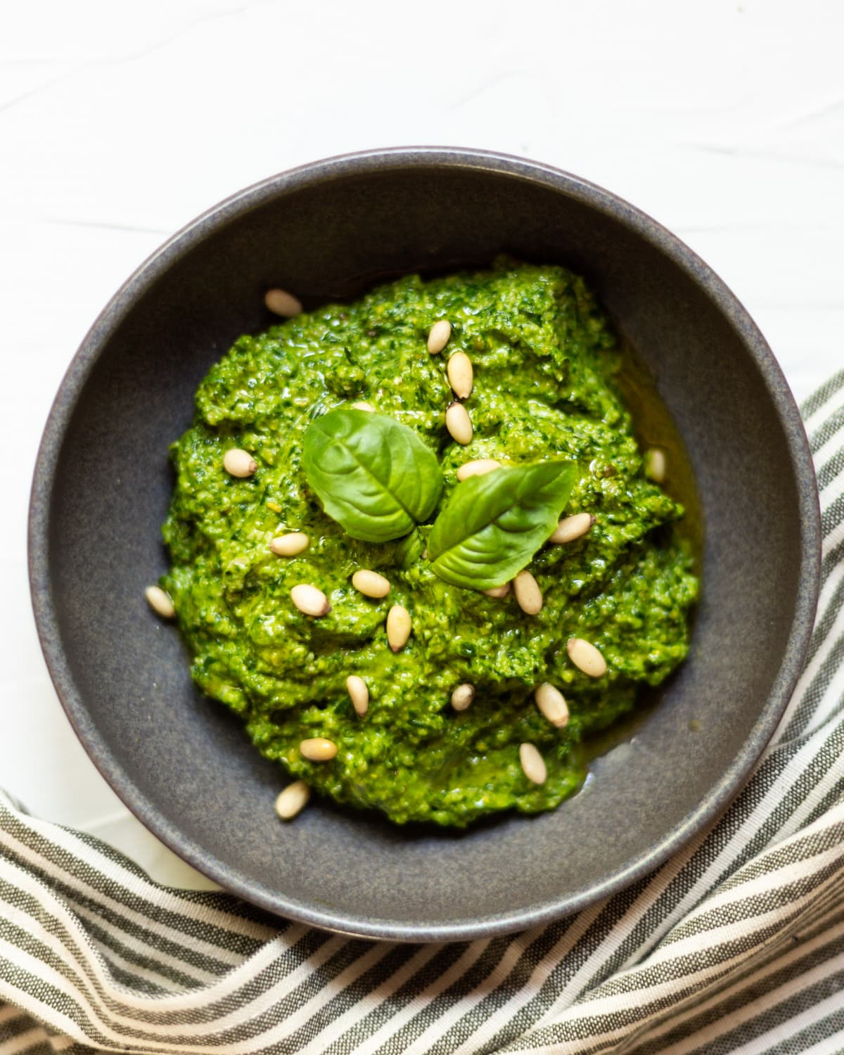 A dark grey bowl filled with basil pesto, topped with pine nuts, fresh basil and olive oil.