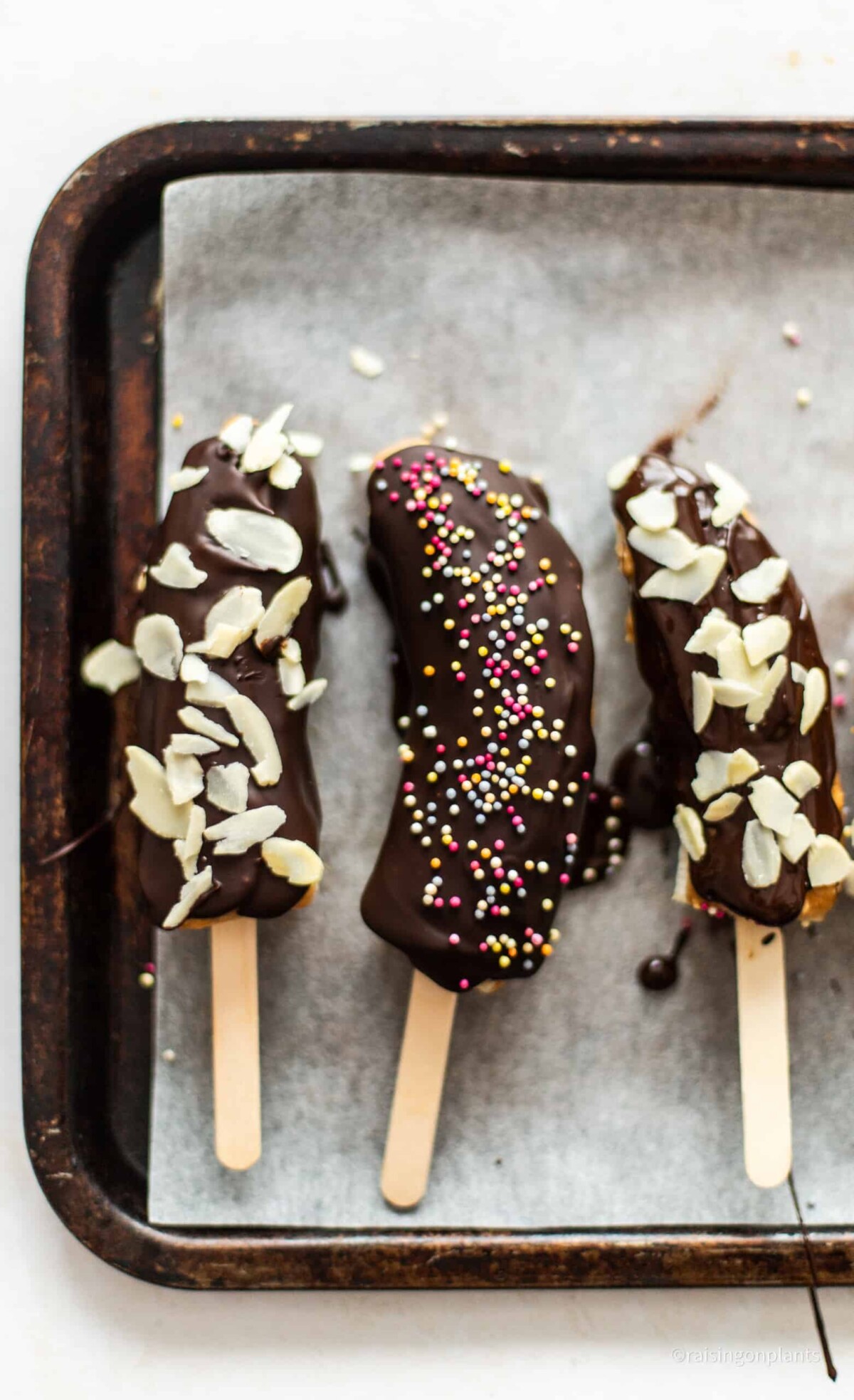 Three chocolate coated banana lollies on a baking tray lined with baking paper.